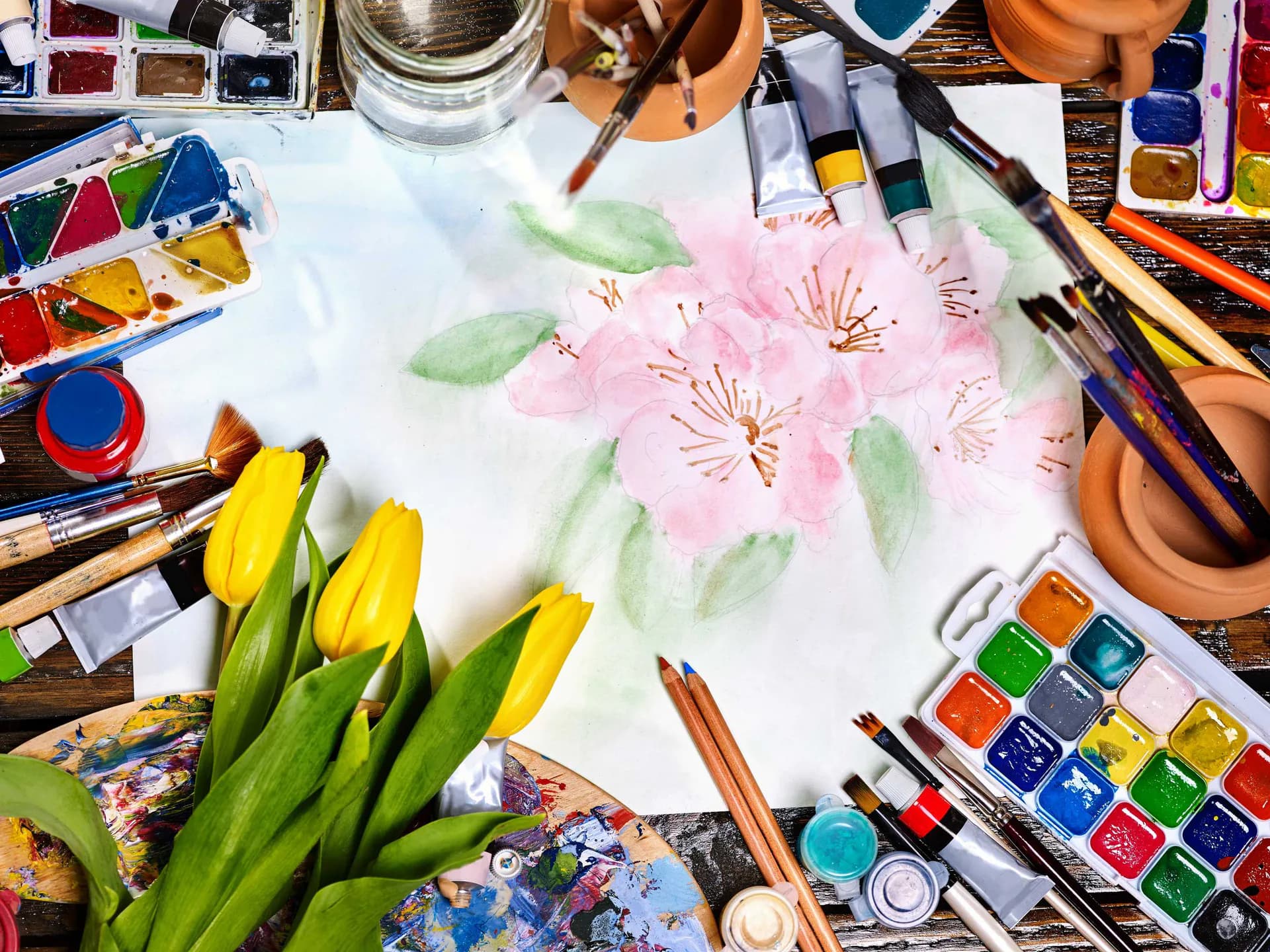 Watercolor painting of pink flowers with green stems and leaves on white paper, surrounded by artist's palette, brushes, and yellow tulips.