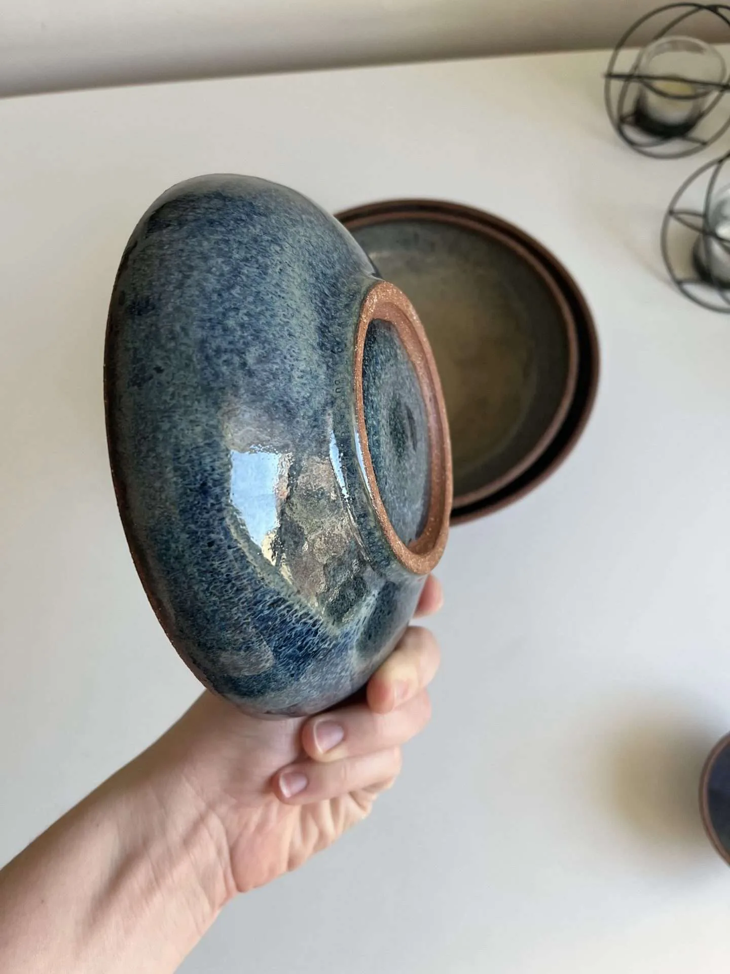 Hand holding an ae stoneware ceramic vessel with glossy teal glaze and spiral texture on base, terracotta rim visible.