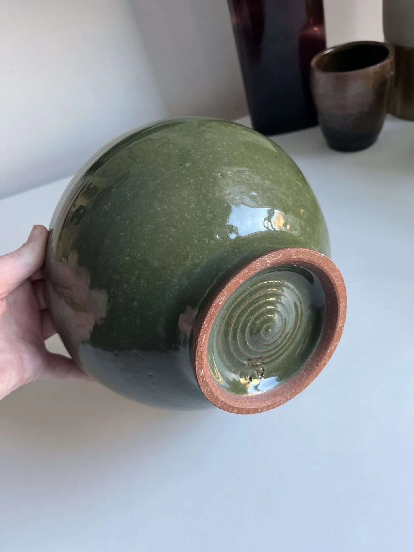 Hand holding ae stoneware spherical ceramic vessel in matte sage green with concentric spiral design on underside base.