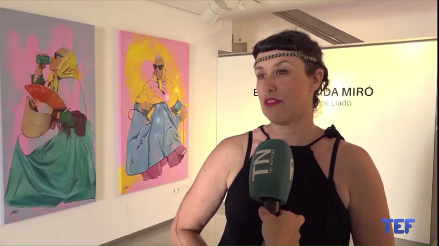 Person wearing black sleeveless top and headband, holding microphone, speaking in front of colorful artwork.