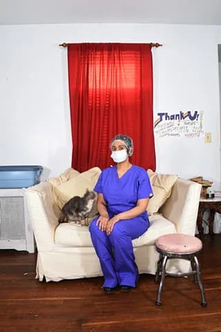 Mixed-media installation by Alicia Grullon: a person in blue scrubs and mask sitting on a cream-colored armchair with a black cat, against white walls with red curtains and handwritten text.