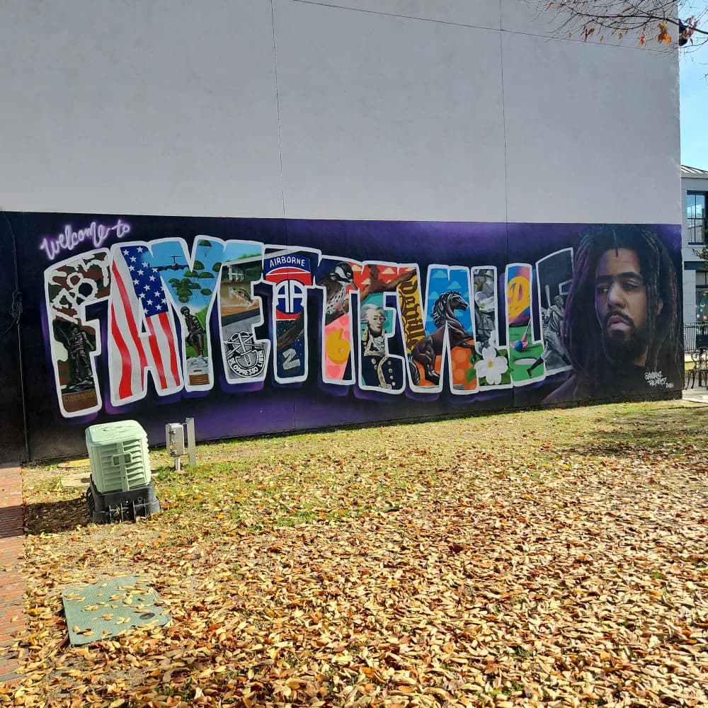 Outdoor mural on white wall with purple base, featuring colorful graffiti lettering and a portrait of a bearded man with dreadlocks on the right.