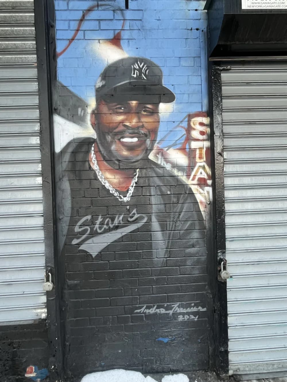 Mural portrait of a smiling man wearing a Yankees cap and gold chain, painted on a dark brick wall with blue background.