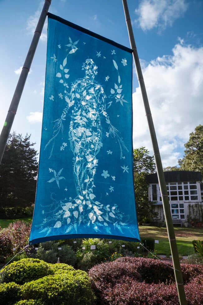 Cyanotype fabric banner depicting a human figure formed from botanical leaves and plants, displayed outdoors on bamboo poles