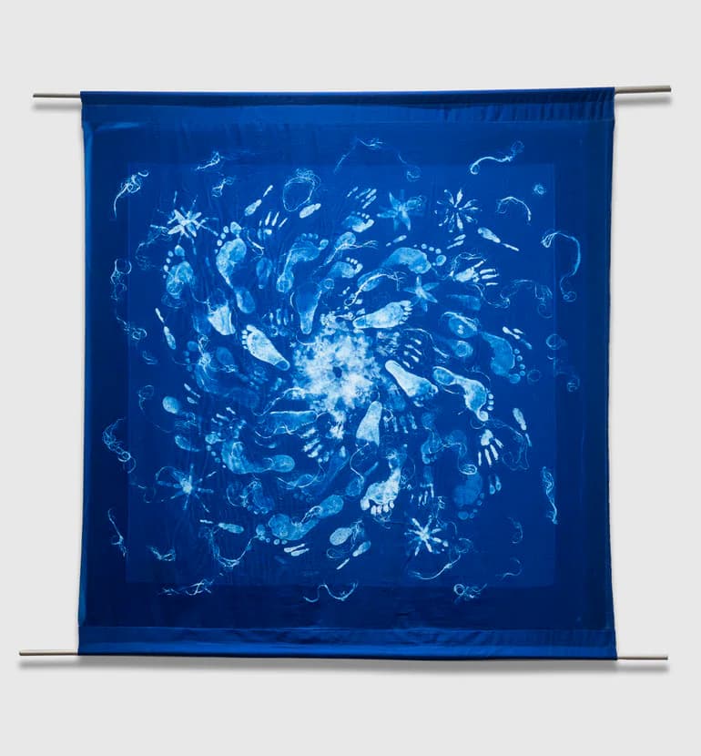 Large square cyanotype fabric tapestry with swirling handprints and footprints radiating from center, mounted on metal rods