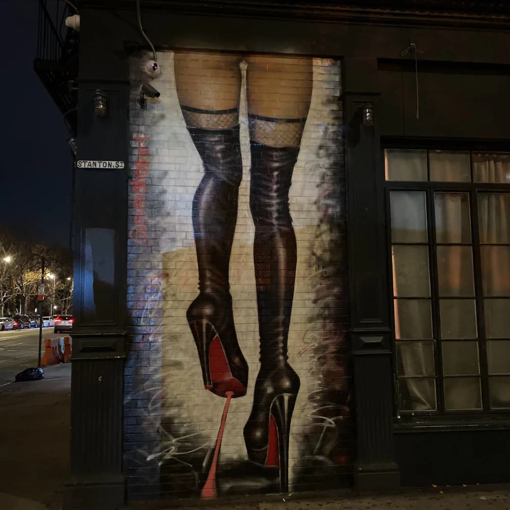Large-scale mural on brick wall depicting legs in black patterned stockings and red-soled heels against cream and tan background, photographed at night.