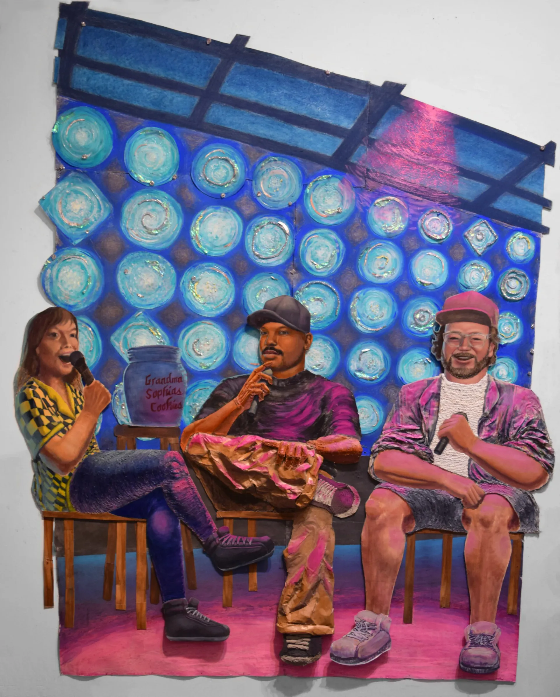 Mixed-media installation: three figures seated on chairs against backdrop of illuminated blue circular objects and magenta-pink floor.