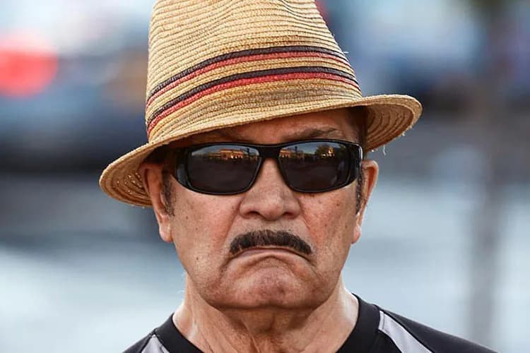 Portrait photograph of a man wearing dark sunglasses and a straw fedora with red and dark striped band, with a distinctive mustache, against a blurred urban background.