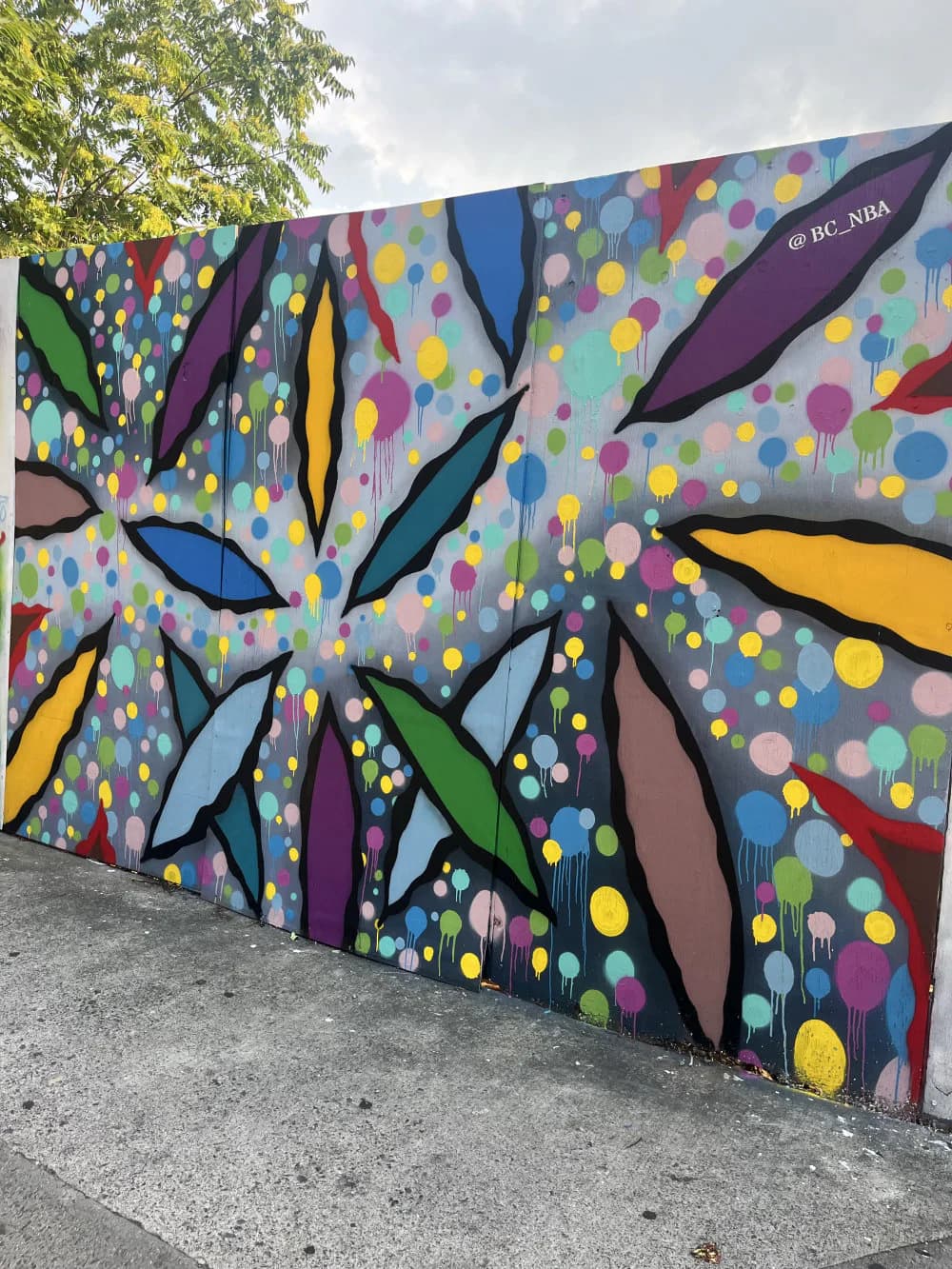 Vibrant mural by BC_nba featuring abstract geometric shapes in black, purple, yellow, green, and blue with colorful circular dots on gray background.