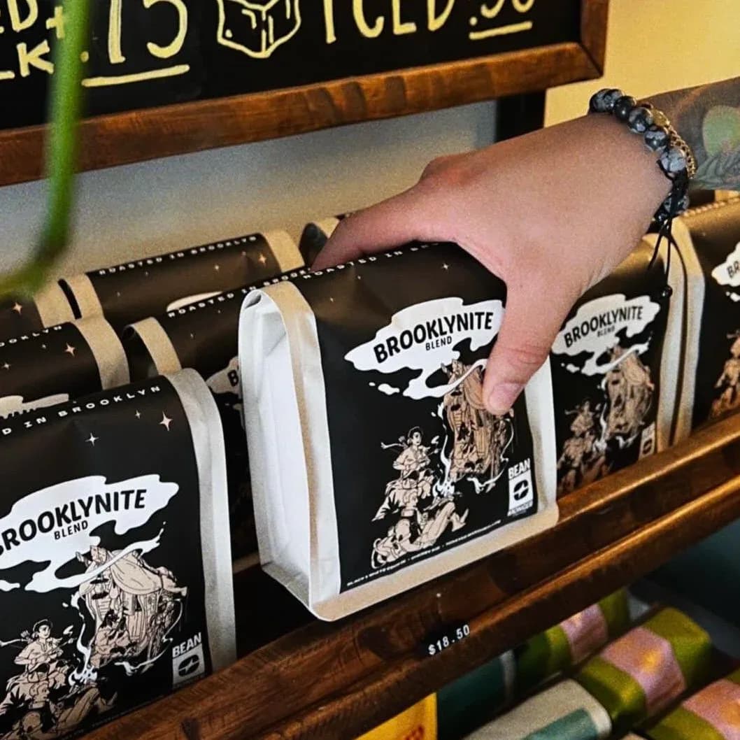 Hand selecting Beanmonger Coffee Brooklynite Blend package from a wooden shelf display with multiple black and cream packages.