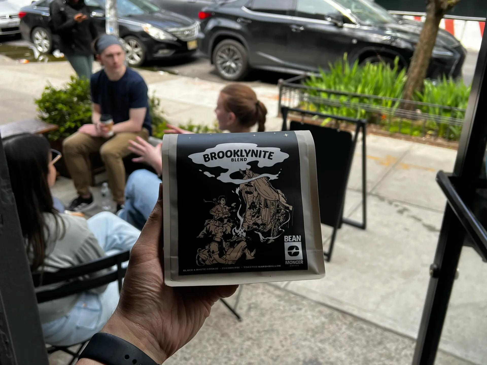 Person holding Beanmonger Coffee Brooklynite Blend package outdoors on a city street with people sitting in background.