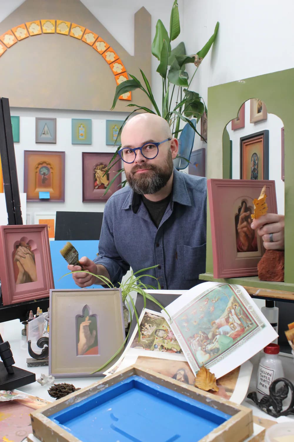 Studio photograph of artist Ben Cowan at desk surrounded by framed small paintings in pink and neutral tones, with reference materials and plants visible.