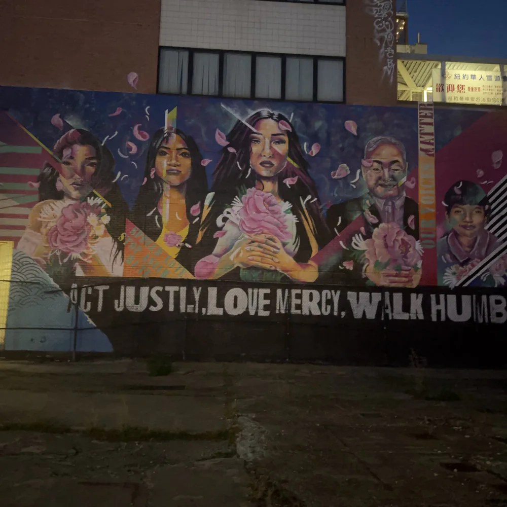 Mural featuring four colorful portrait figures holding pink flowers against a dark background with text reading 'JUST JUSTIN, LOVE MERCY, WALK HUMBLY'.