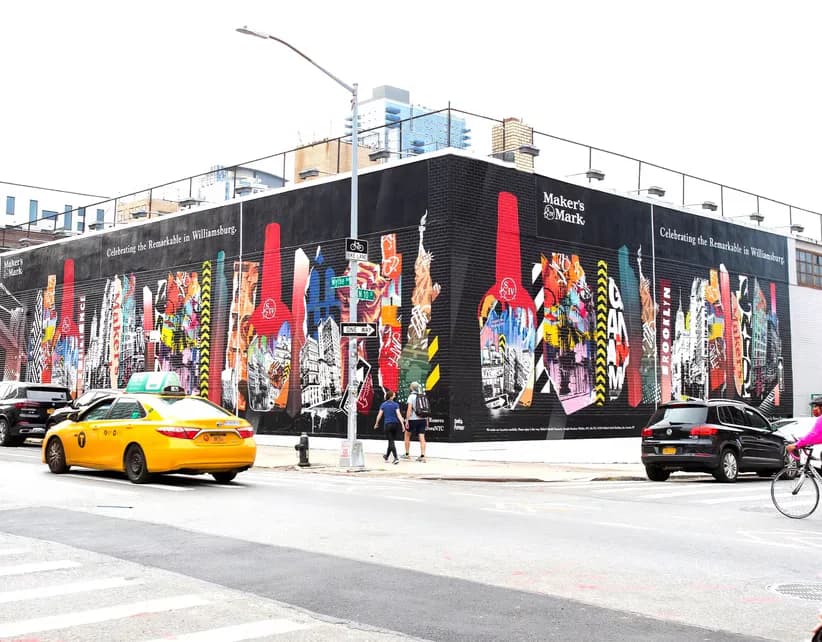 Photograph of a large black building corner covered in vibrant Bianca Romero murals with colorful figures, street level with yellow car parked nearby.