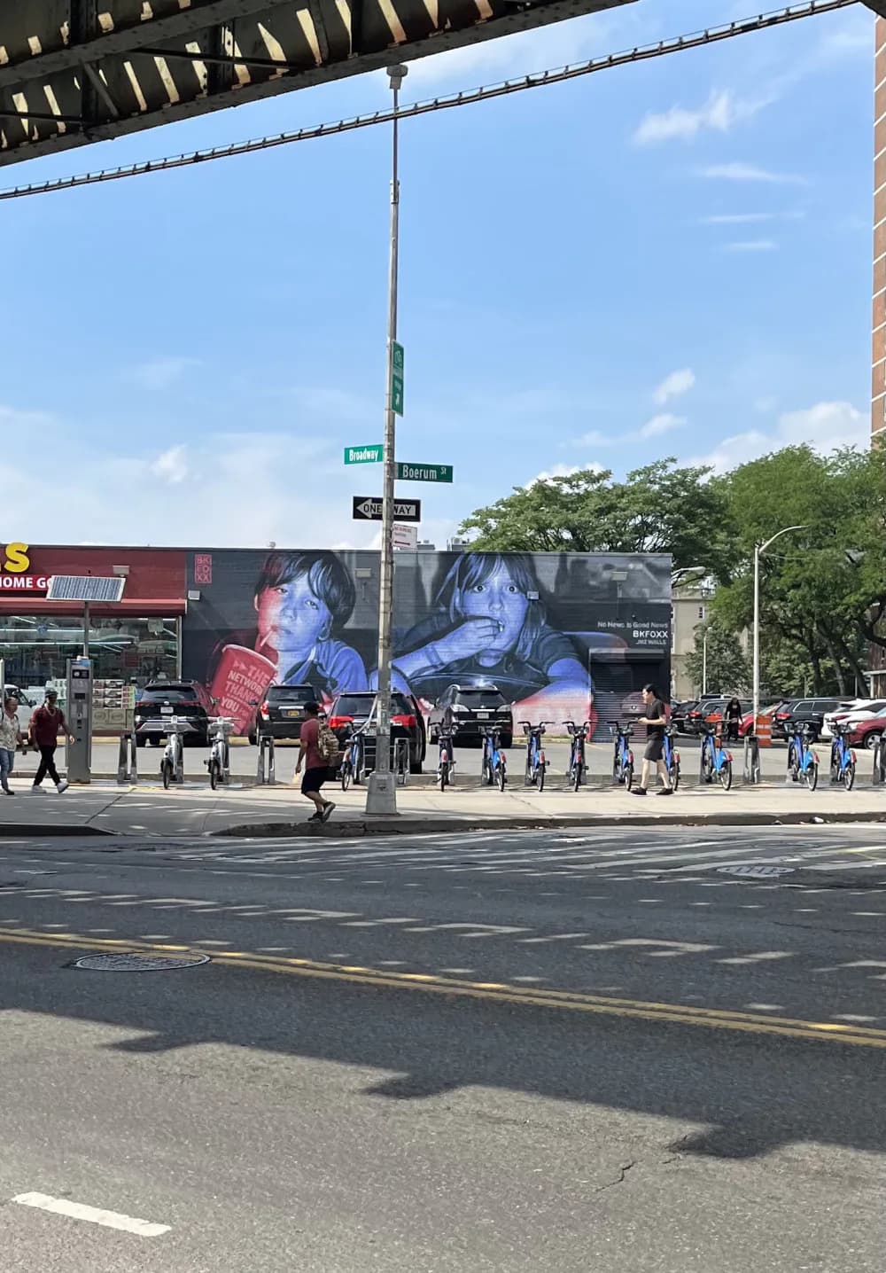 Large-scale outdoor mural depicting two portraits in blue and red tones on a building facade in an urban plaza.