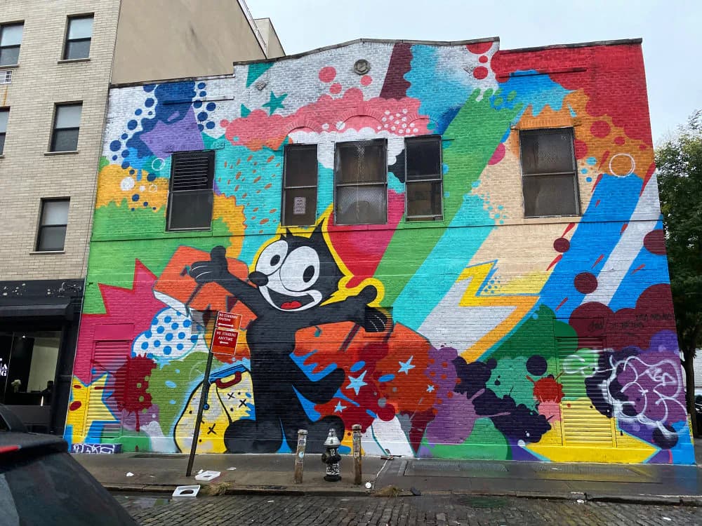 Vibrant street mural by BudaReal featuring a cartoonish black character with white gloves dancing among colorful abstract shapes, geometric patterns, and playful design elements in red, blue, green, yellow, and purple.