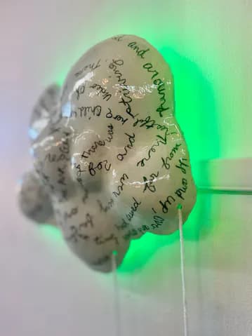 Organic brain-shaped sculpture with handwritten text covering its surface, illuminated by bright green neon light on one side.