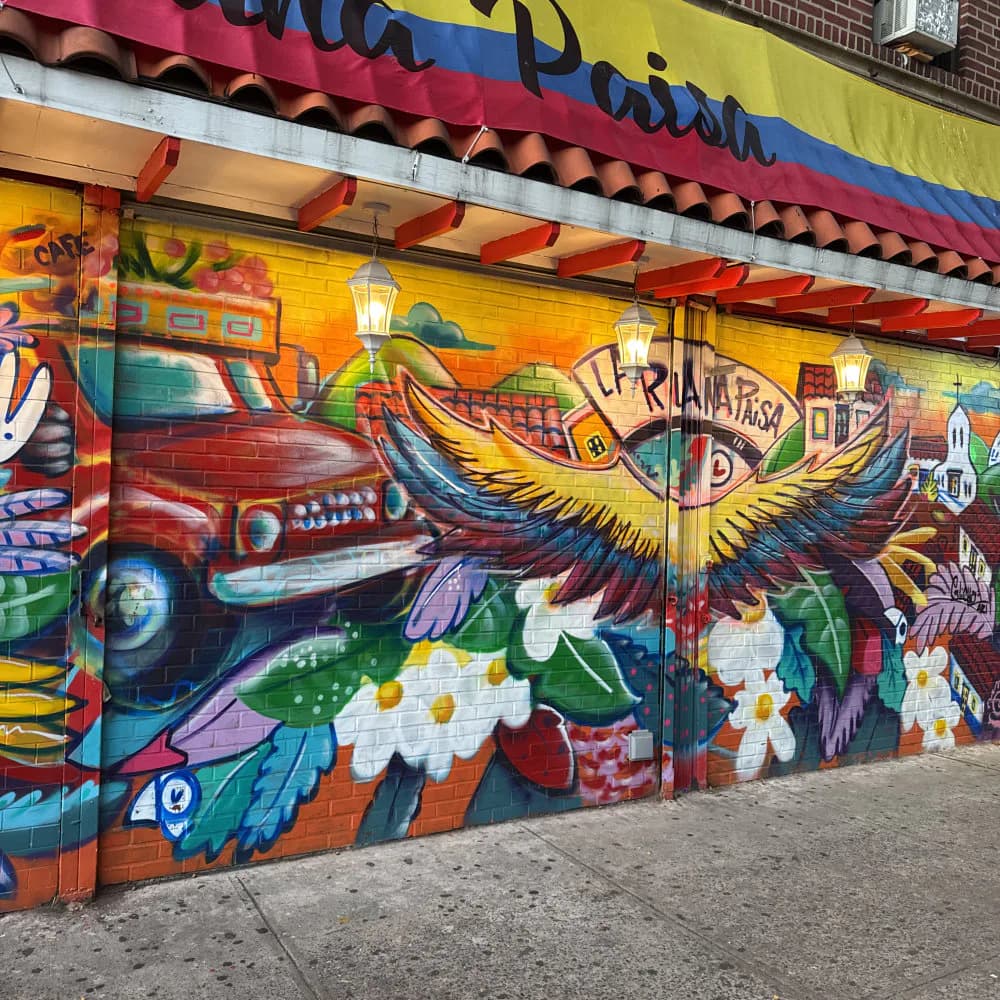 Vibrant street mural featuring a winged eye motif in gold and purple, surrounded by colorful abstract elements and flowers on a brick wall.