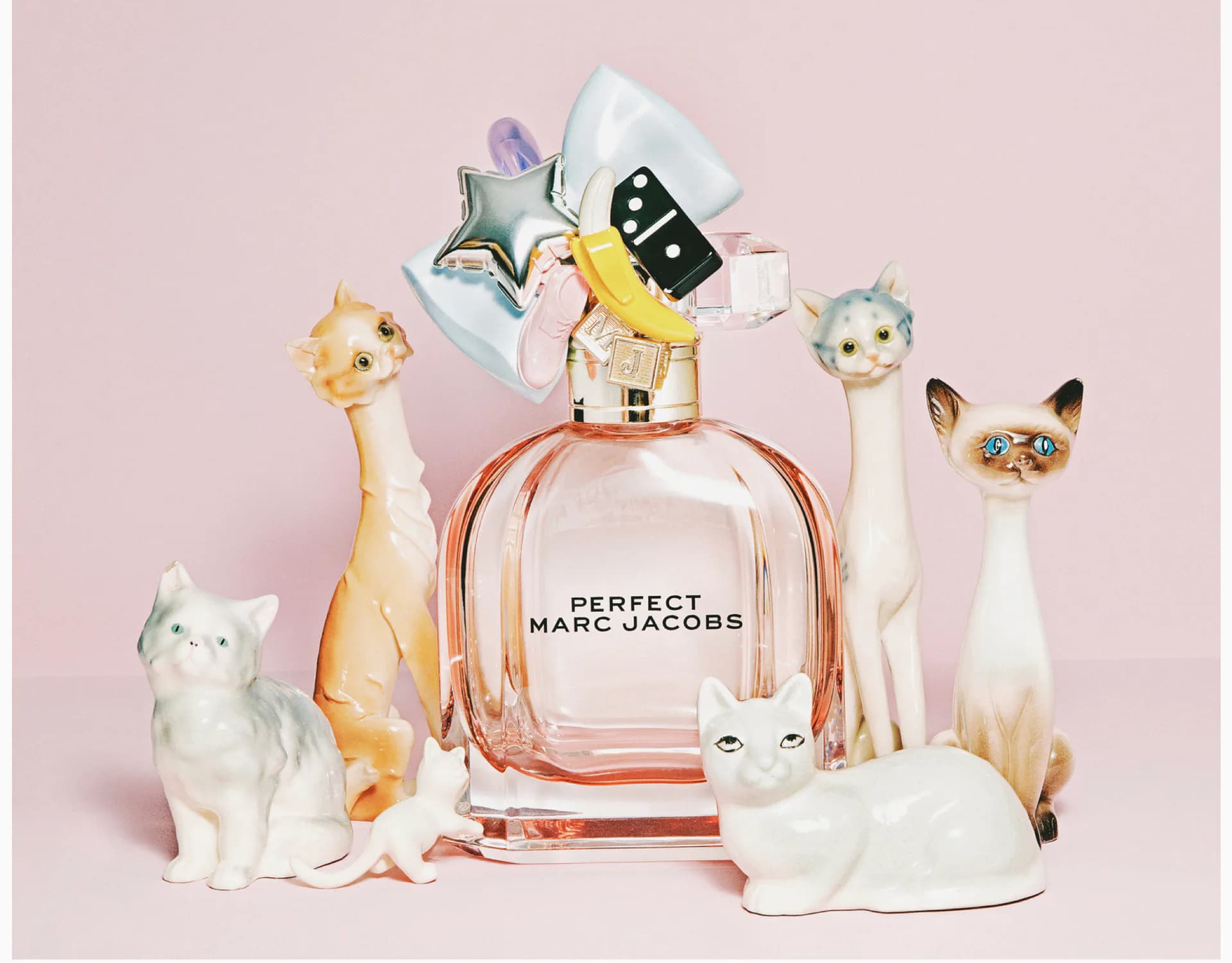 Product styling photograph of Marc Jacobs 'Perfect' perfume bottle surrounded by colorful ceramic cat figurines and decorative objects on pink background.