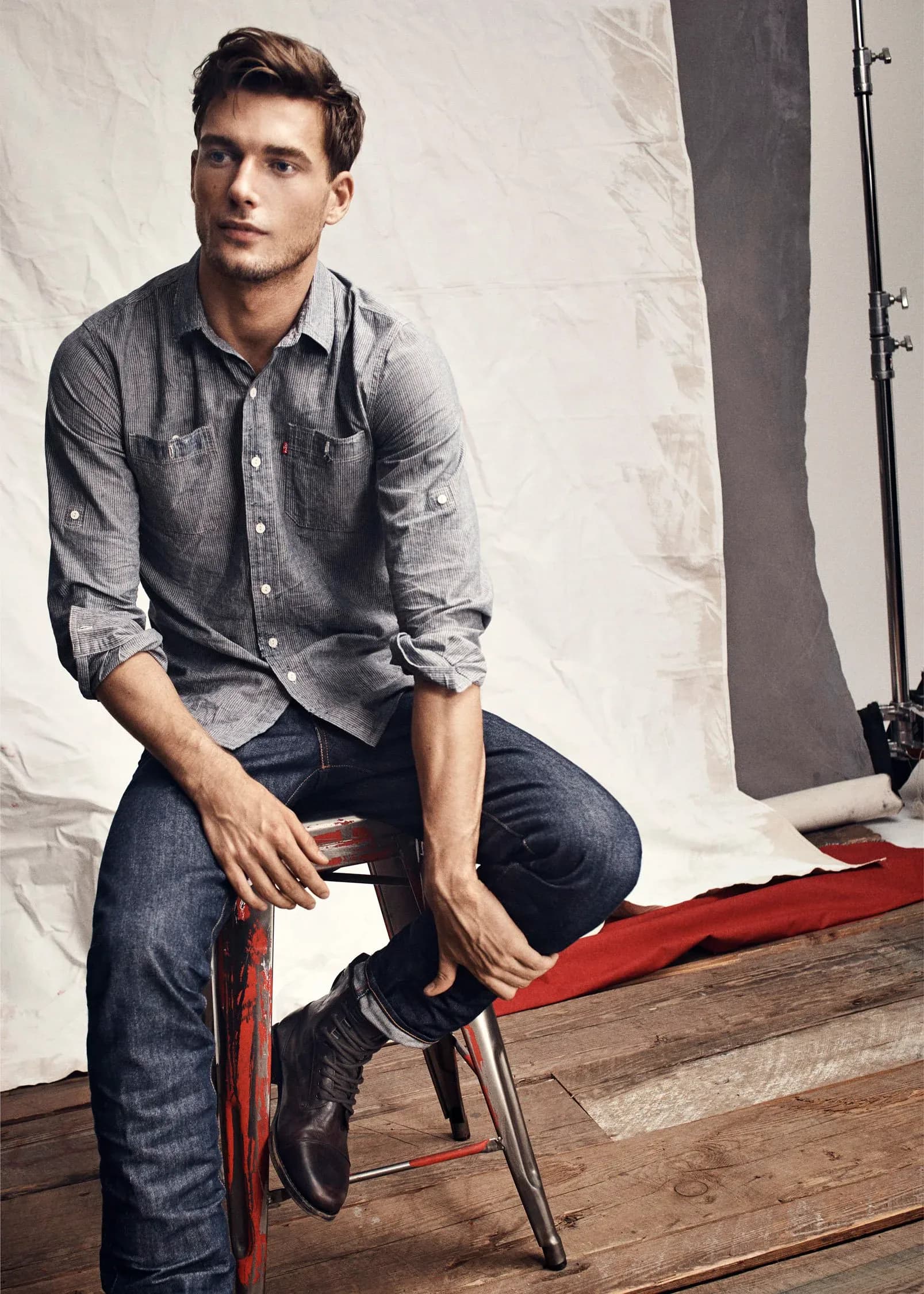 Styled fashion portrait of a man in denim shirt and dark shoes seated on red stool against white fabric backdrop with gray fabric panel and lighting equipment.