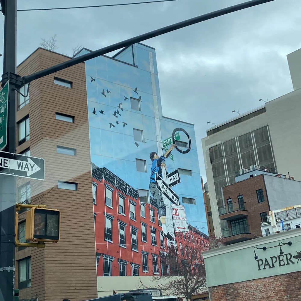 Street corner with mural artwork on building facade featuring figure on bicycle and flying birds against blue sky background.