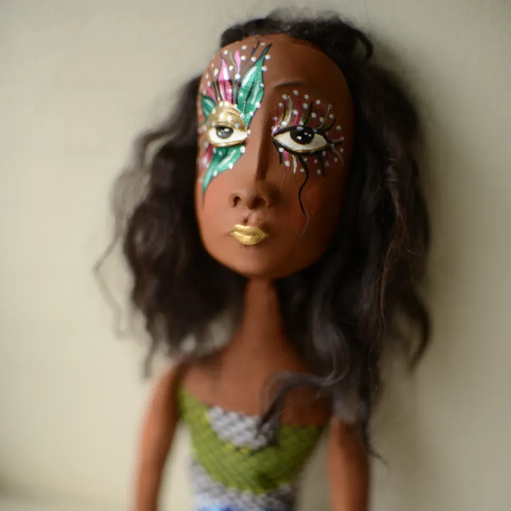 Sculptural doll head with long dark hair, decorative face paint in pink, green, and white, and gold lips. Polymer sculpture.