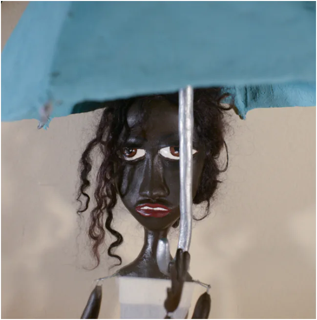 Sculptural doll with dark face, long black curly hair, holding a blue umbrella. Mixed media sculpture with painted details.