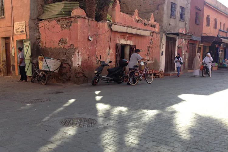Street scene in a weathered terracotta-colored medina with vehicles, pedestrians, and dramatic shadows cast across the ground by sunlight.