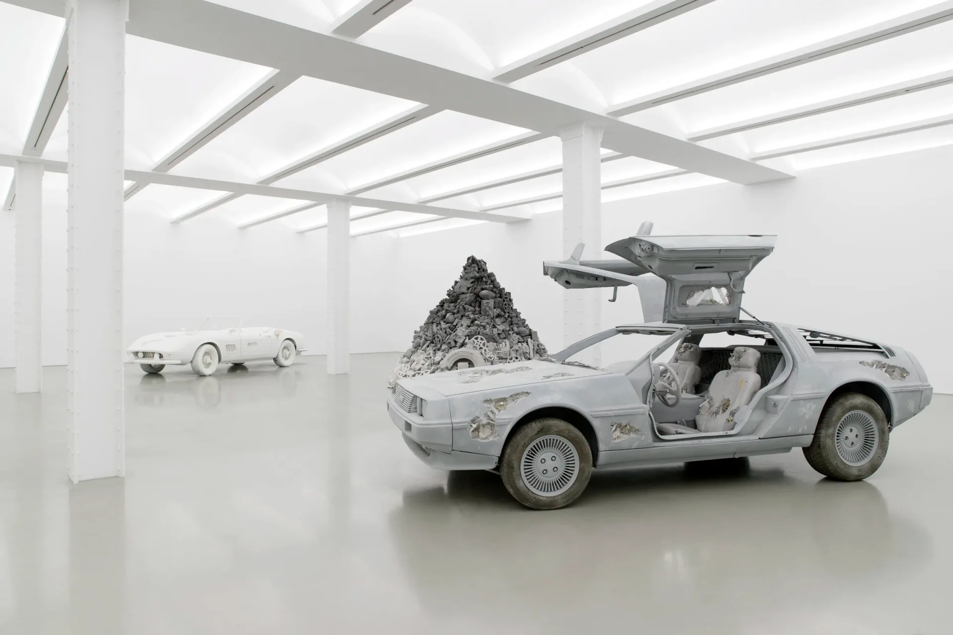 White gallery space with minimalist sculptural installation featuring a deconstructed white sports car, small pyramid form, and toy vehicles rendered in monochromatic material.