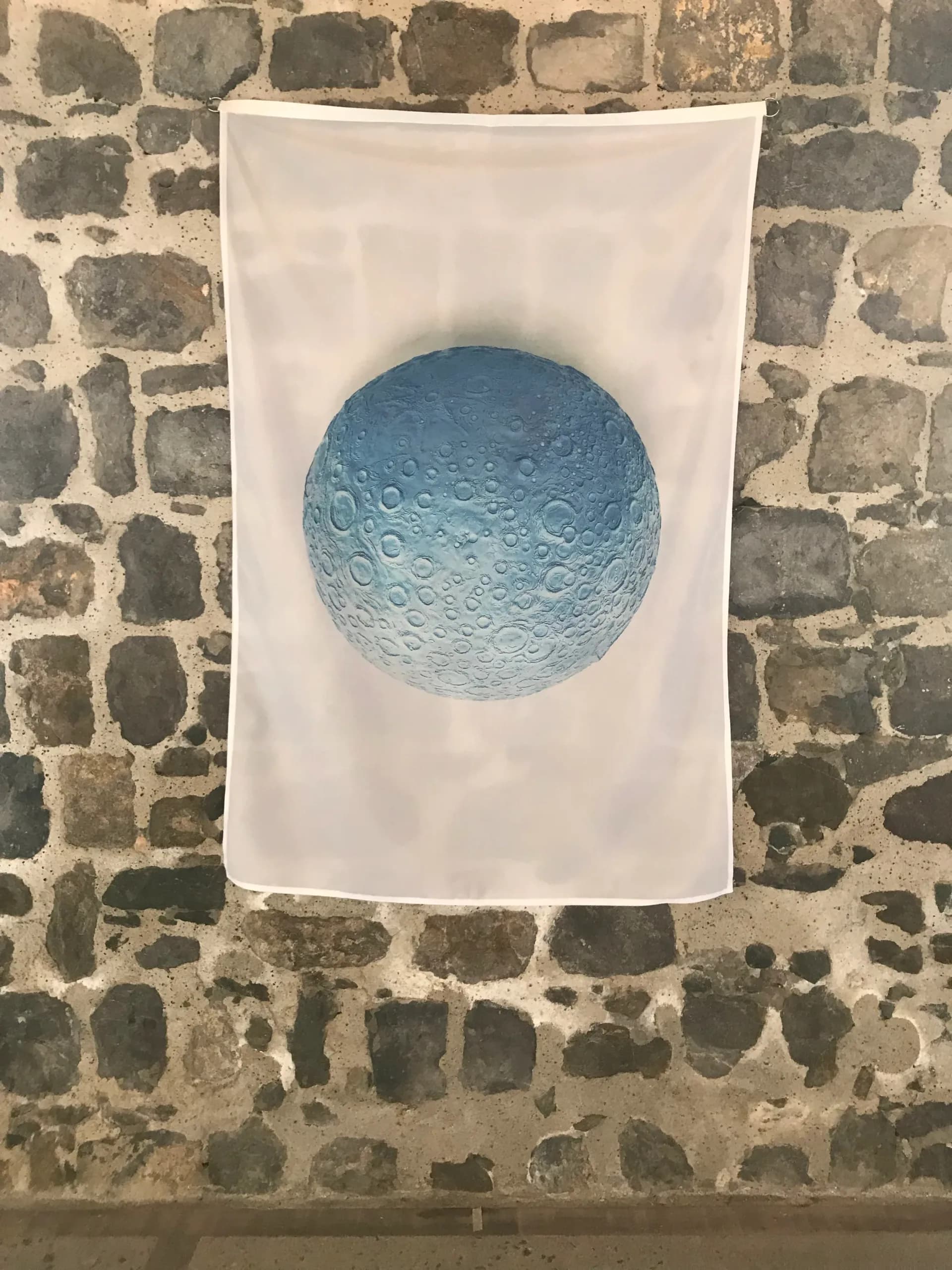 Blue ceramic or cast moon sphere mounted on white fabric banner displayed against weathered stone wall; sculptural celestial reference work.