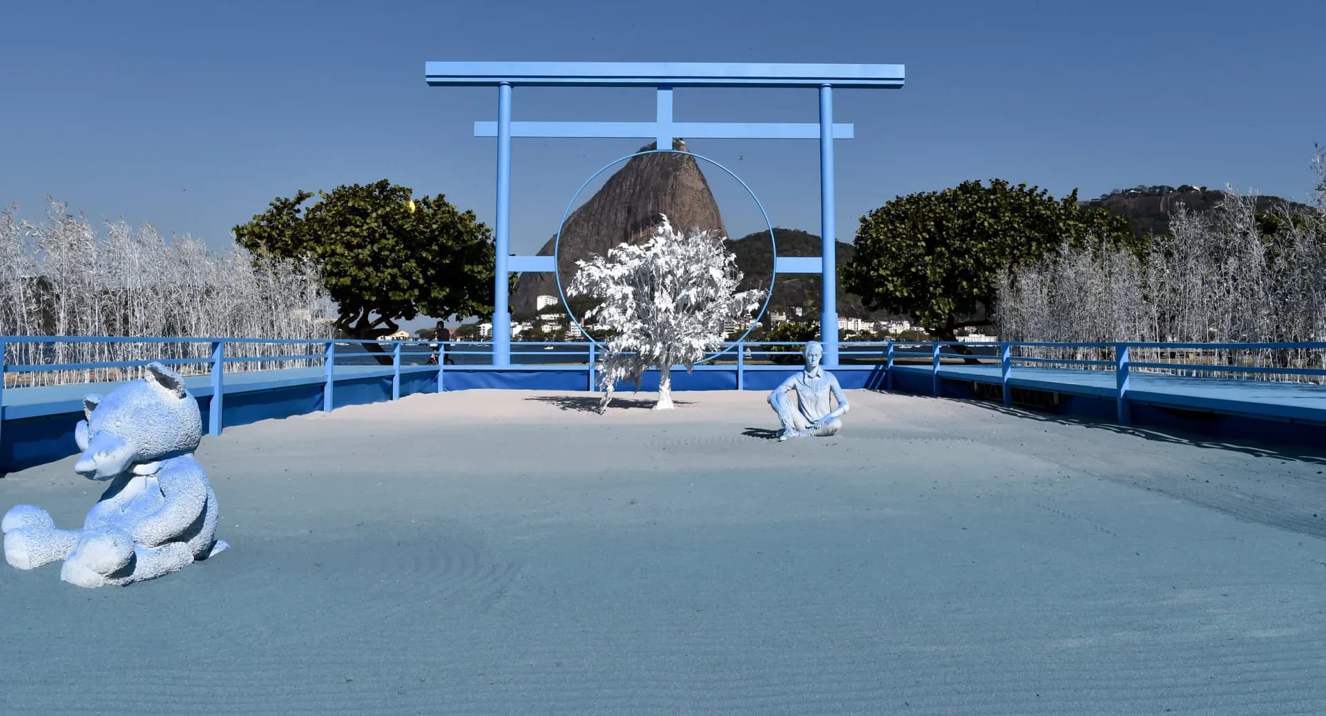 Surreal outdoor installation with pale blue platform, architectural frame, and sculptural forms set against natural landscape with trees and water.