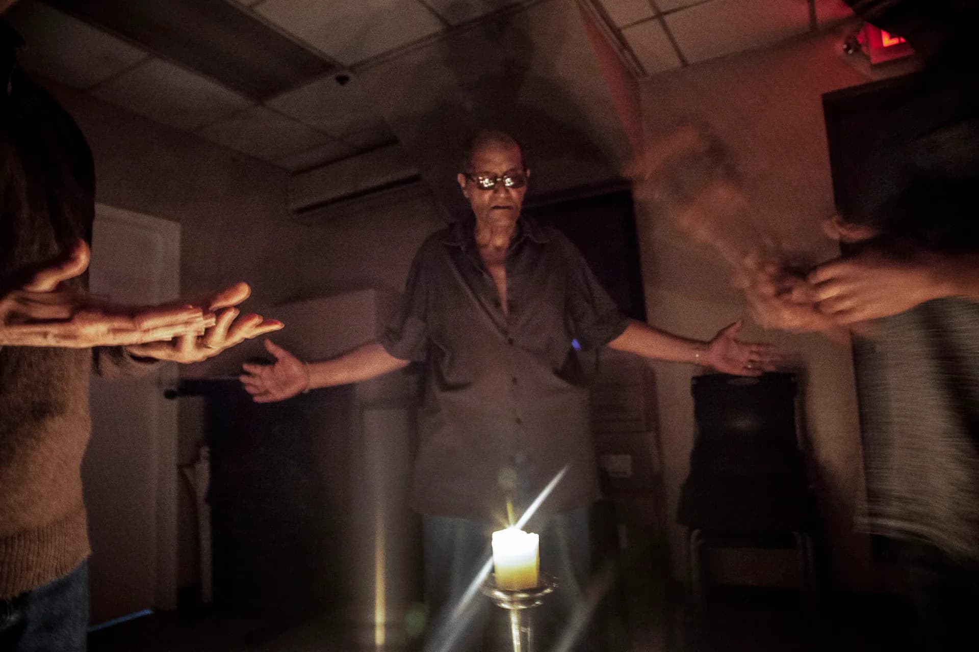 Installation or performance artwork with multiple figures in dark lighting, one central figure in glasses with arms outstretched, holding a glowing candle.