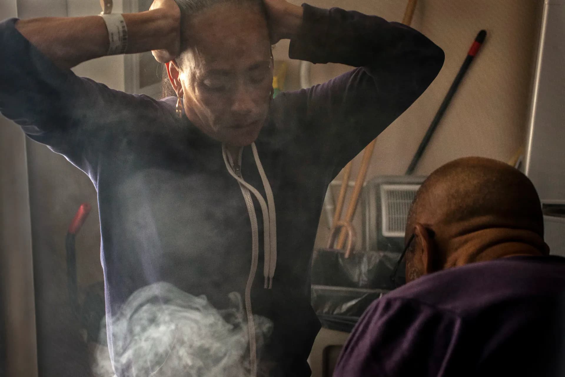 Performance or installation artwork showing figures with smoke or incense, one person with hands raised to head, another seated, in warm indoor lighting.