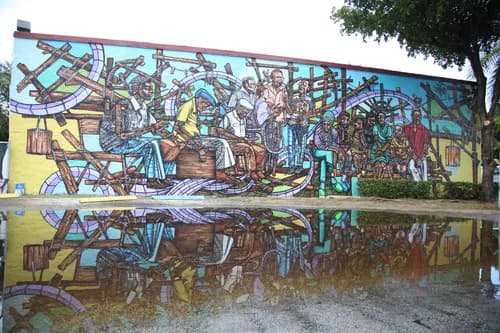 Large-scale mural depicting figures in a community scene with geometric stained-glass style, reflected in a puddle