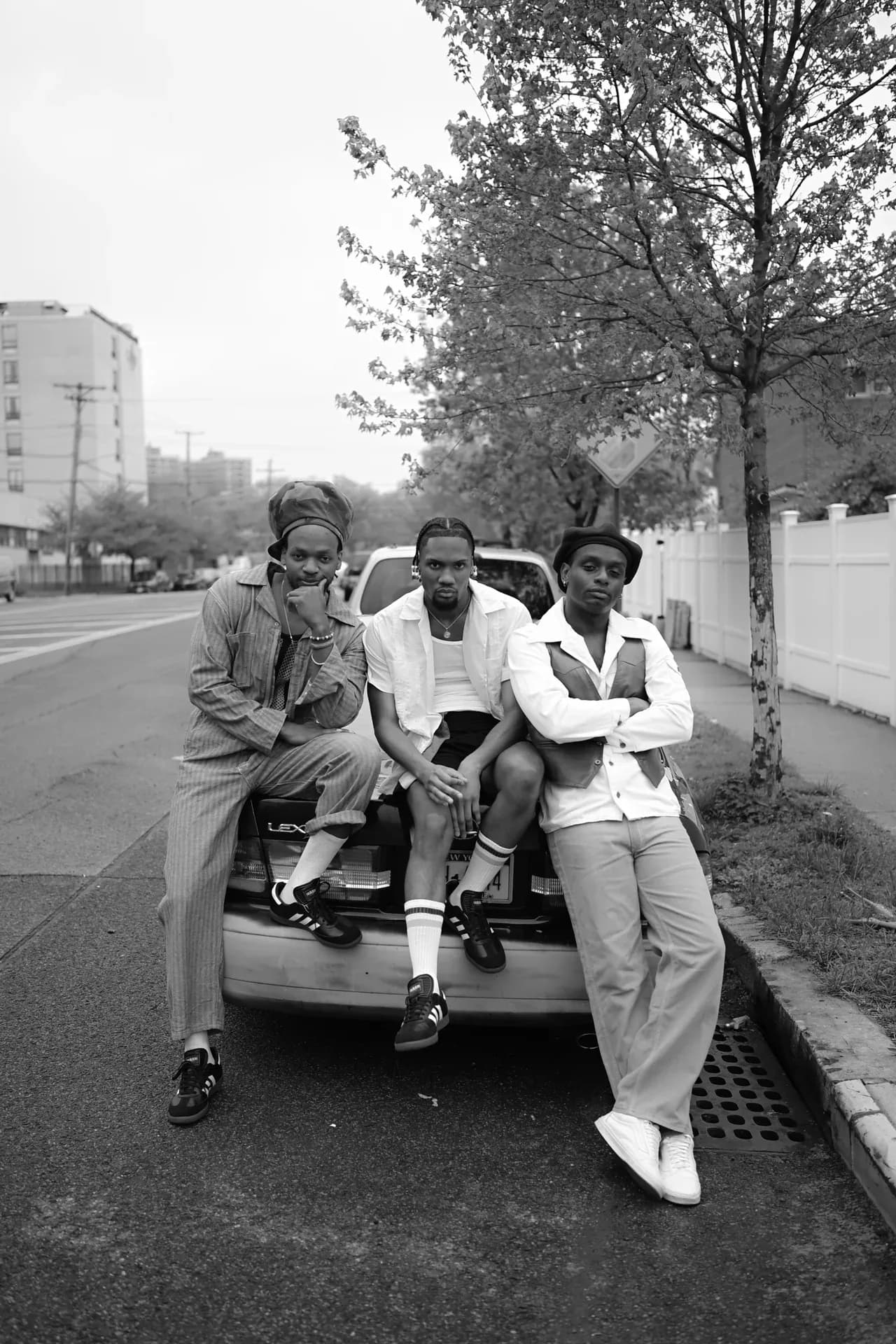 Black-and-white photograph of three stylishly dressed people seated on car hood against urban street backdrop.