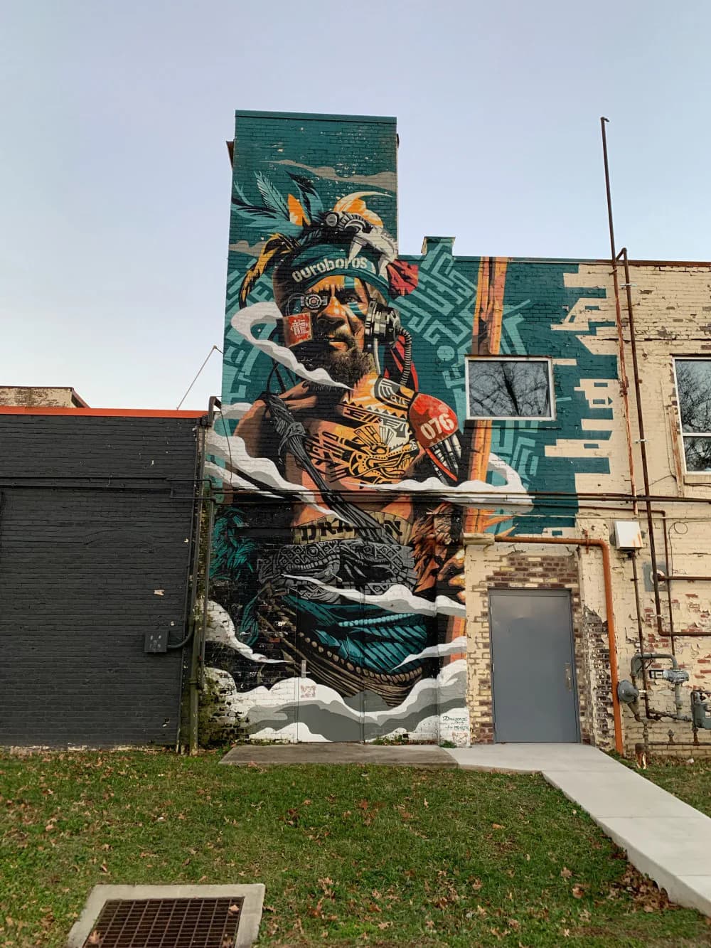 Tall mural featuring stacked figures and creatures in teal, orange, and cream on building wall with geometric patterns and wildlife elements.