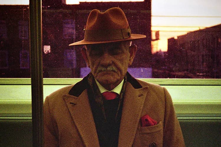 Man in camel coat and burgundy fedora standing in front of illuminated storefront at dusk, shot in warm and cool color tones.