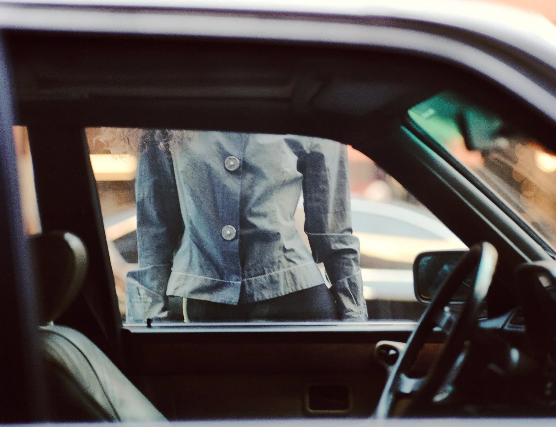 Partial view through car window of person in denim jacket; film photography with vintage color treatment and voyeuristic composition.