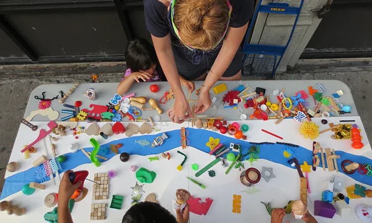 Overhead view of collaborative mixed-media installation with colorful objects, craft materials, and artistic elements arranged on white surface.