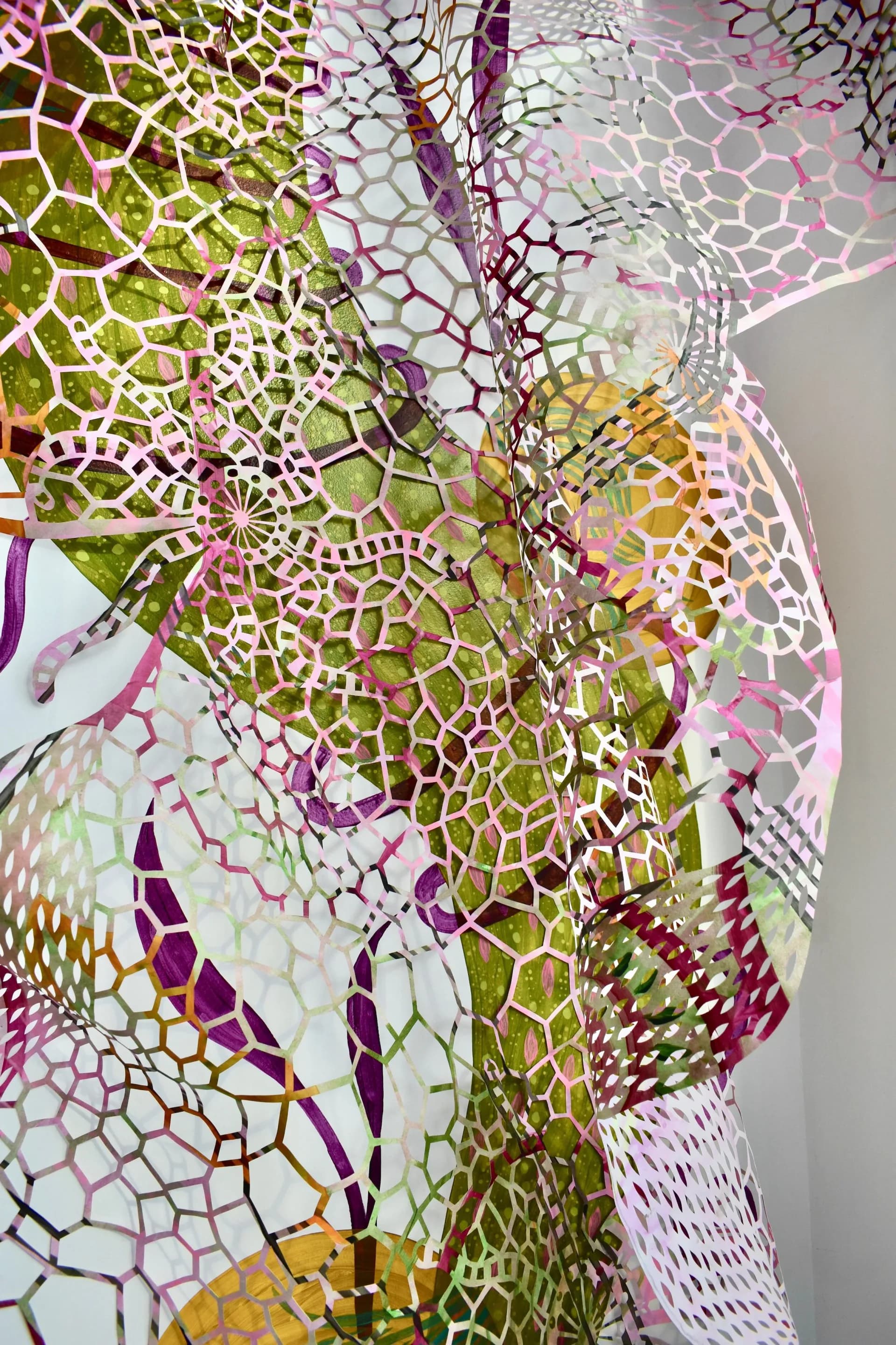 Large-scale mixed-media installation detail: layered transparent and translucent fabrics in magenta, lime, and cream with geometric cutout patterns creating dimensional overlap.