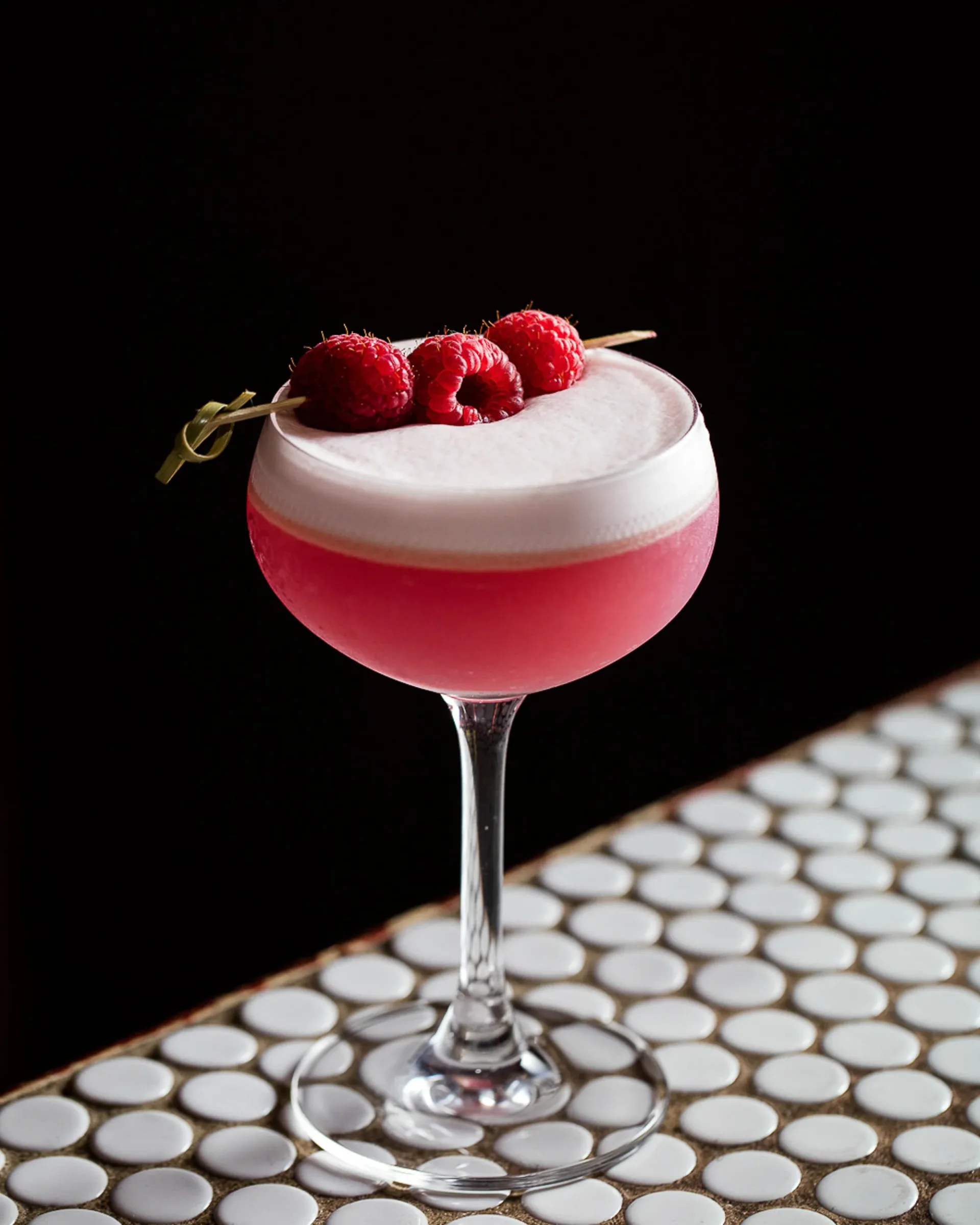 Raspberry sour cocktail with foam and fresh raspberries in a coupe glass against black background