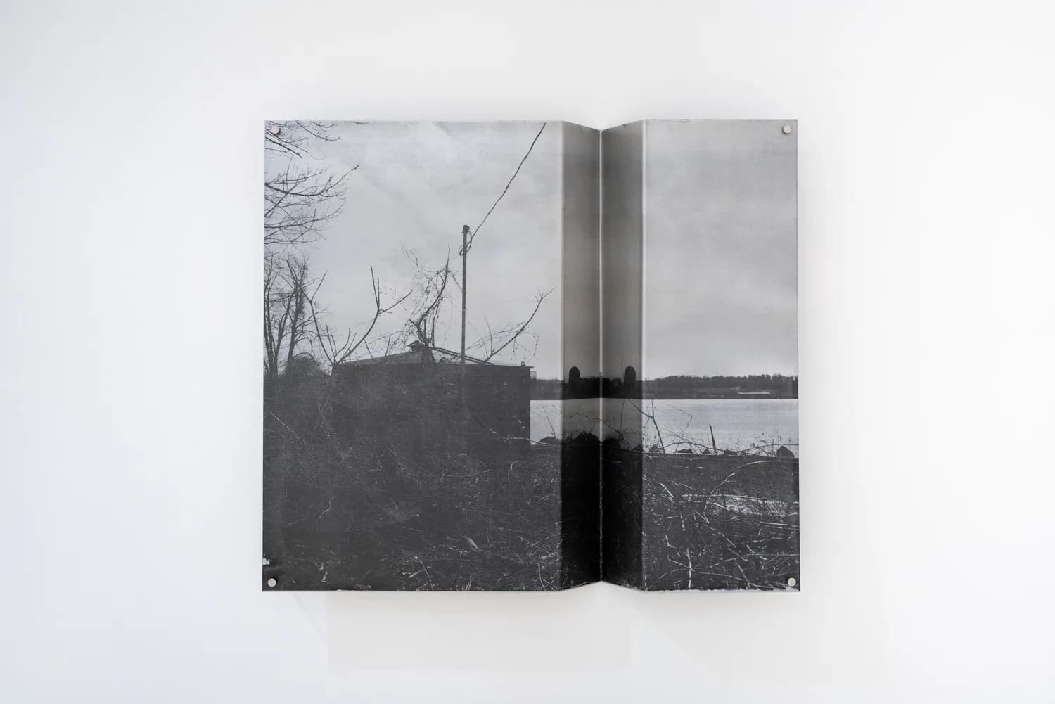 Folded black and white photographic print mounted on wall with rivets, depicting industrial landscape with trees and water