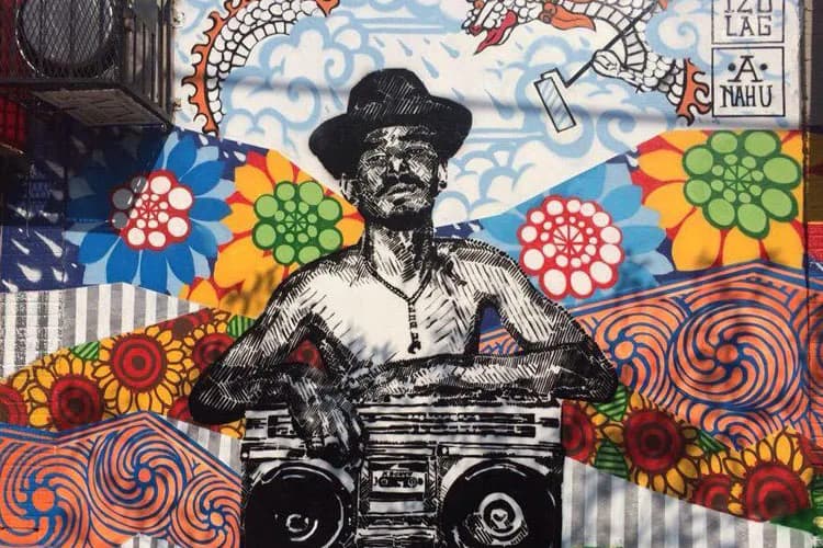 Mixed-media mural by FACES FROM THE BLOCK featuring a black-and-white portrait of a man in a hat holding a boombox, surrounded by vibrant colorful flowers, spirals, and geometric patterns on a striped background.