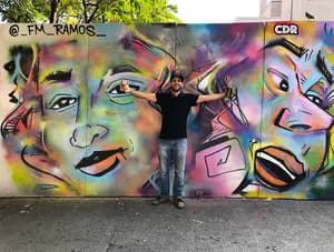 Fausto Manuel Ramos posing in front of vibrant mural featuring three expressive faces with neon colors, painterly strokes, and mixed-media technique.