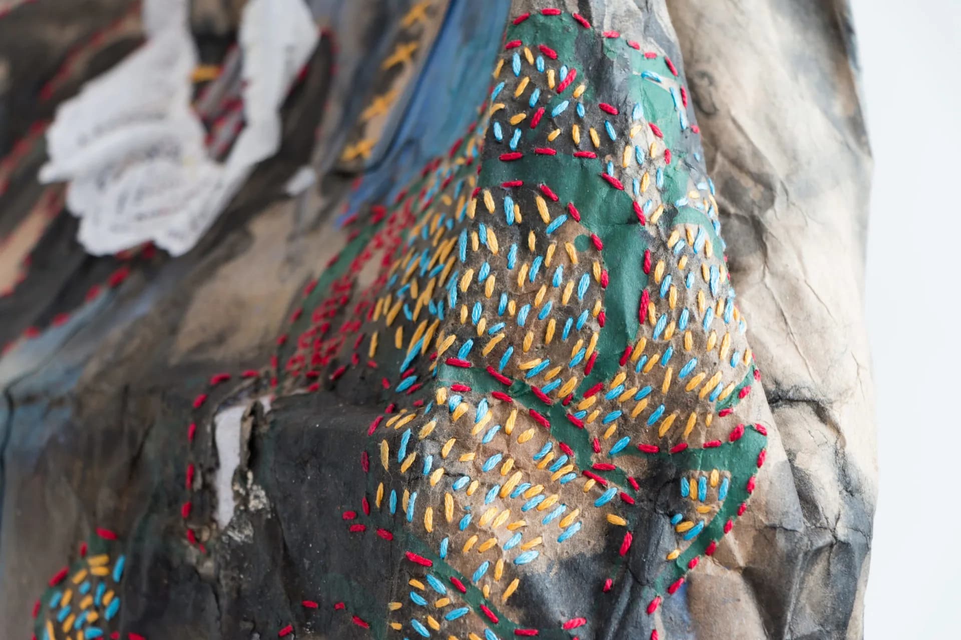 Close-up of mixed-media textile artwork with embroidered or stitched multicolored geometric patterns (orange, green, red, yellow) applied over a painted fabric base in blue and gray tones.