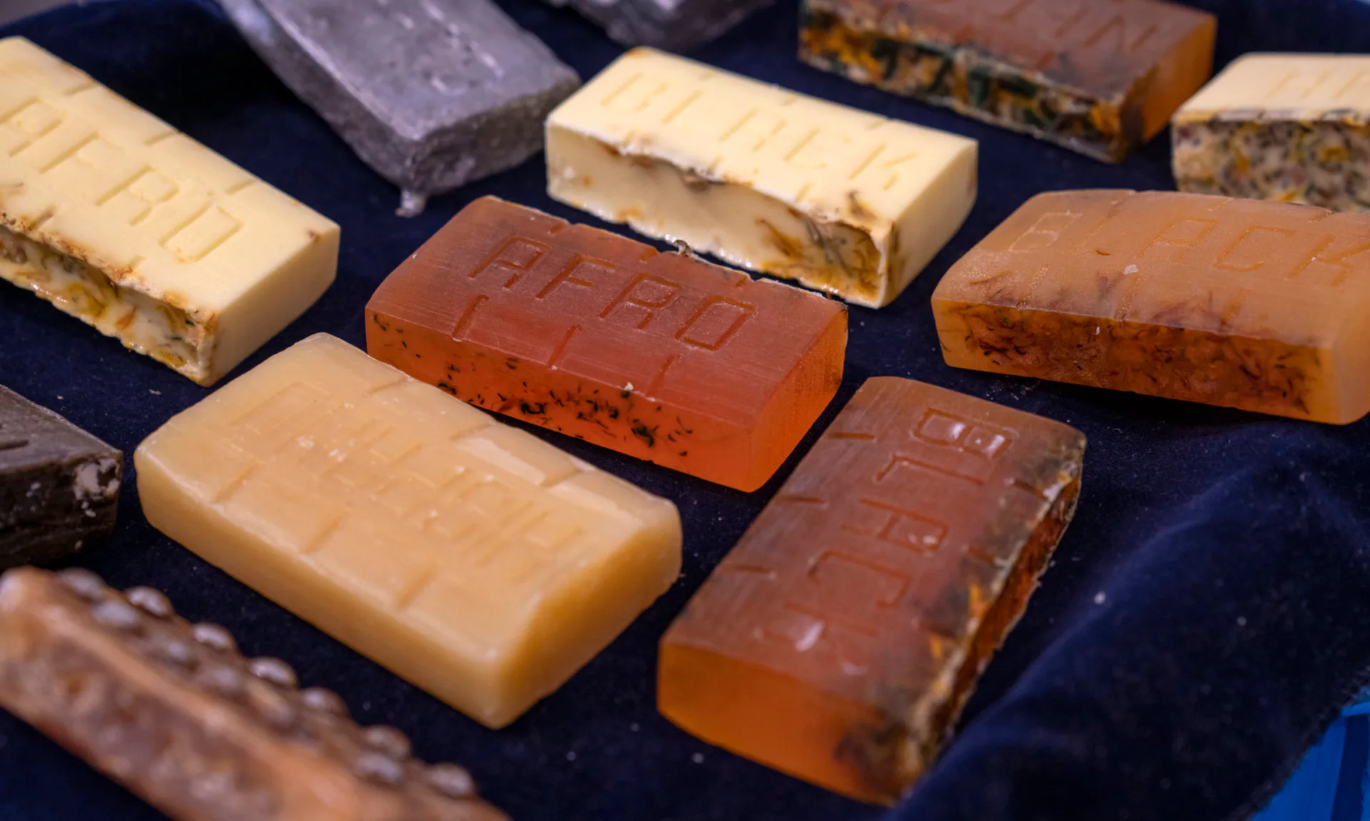 Collection of handmade soap bars in various colors—cream, tan, brown, burgundy, and purple—arranged on a dark surface, some with embossed patterns.
