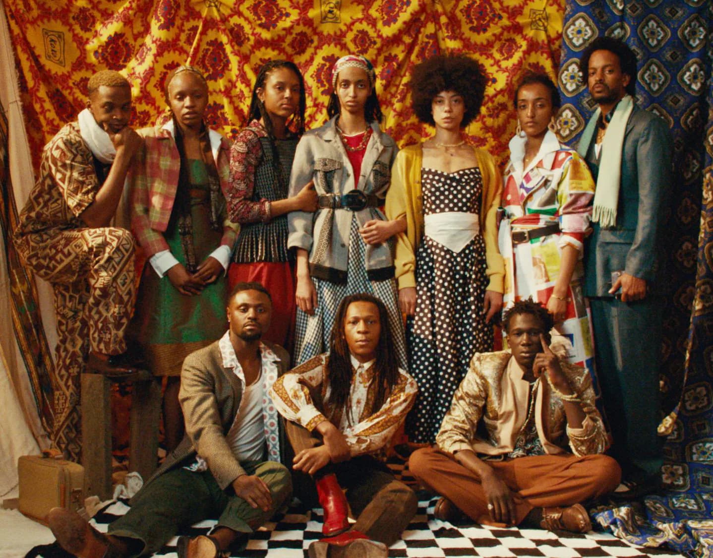 Group portrait of ten people in eclectic mixed-pattern clothing against vibrant patterned fabric backdrop on checkered floor