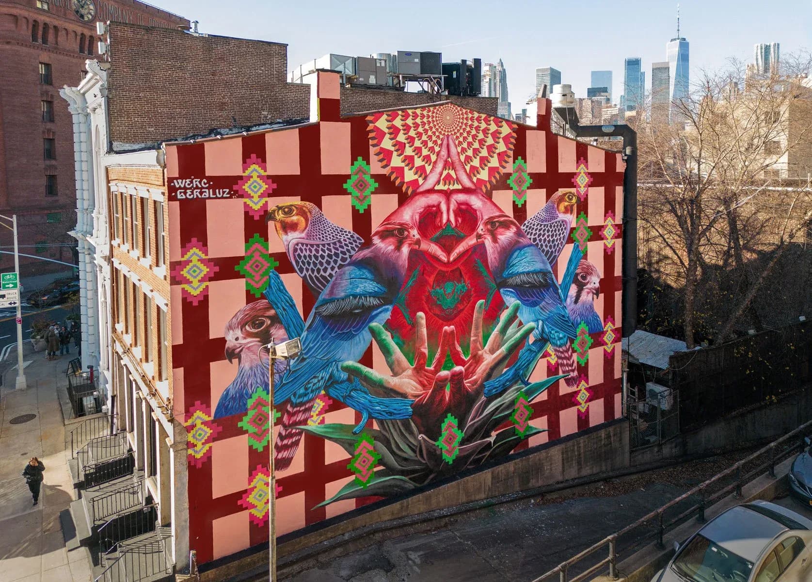Large-scale mural on building corner featuring falcons, hands, and geometric patterns with NYC skyline in background