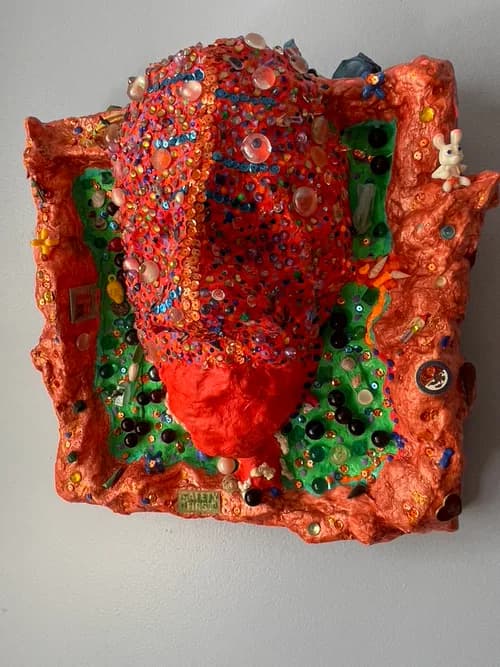 Sculptural mixed-media face in orange with embedded sequins, beads, toys, and small objects
