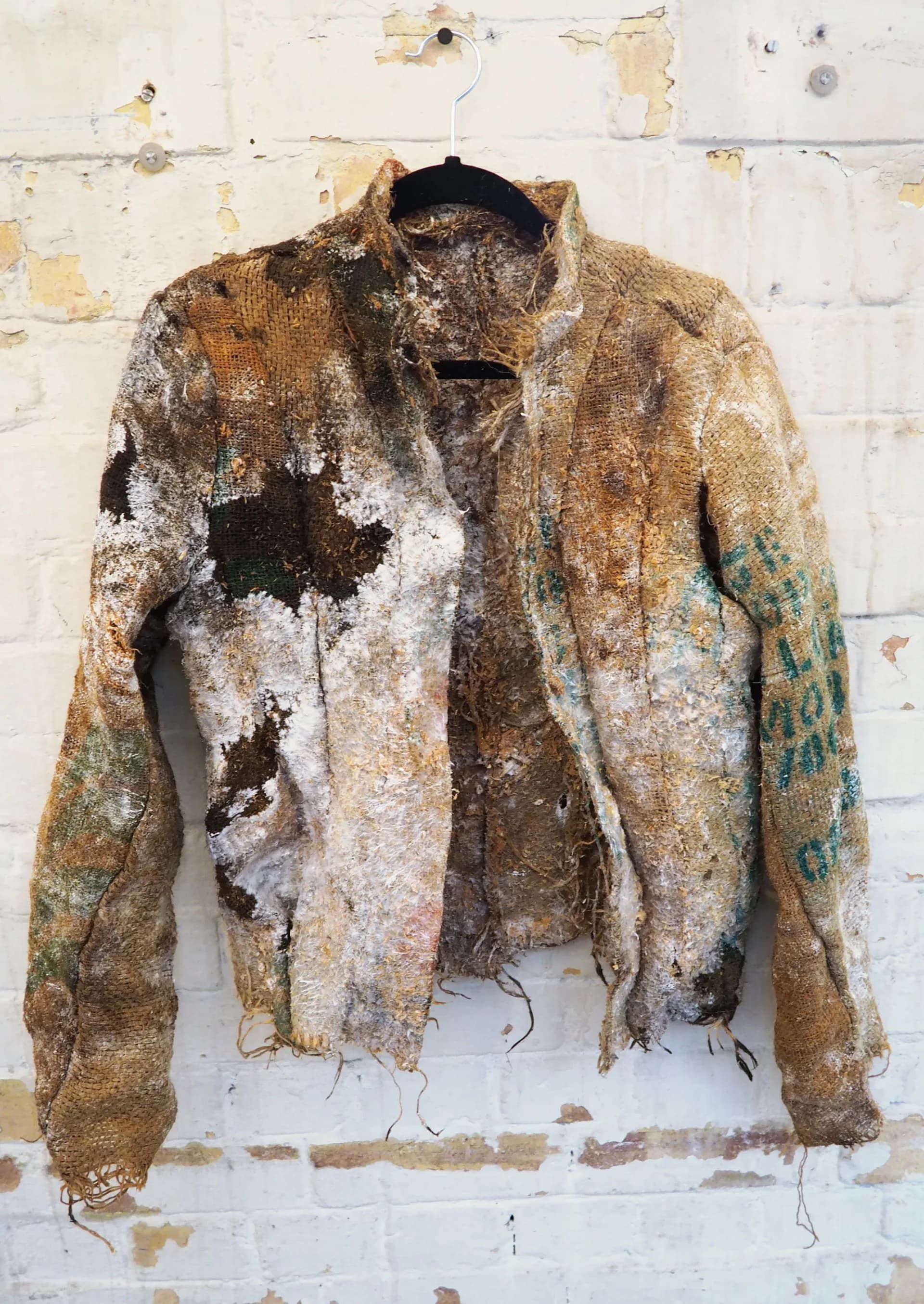 Hessian jacket colonized by mycelium and decomposed materials hung on white brick wall